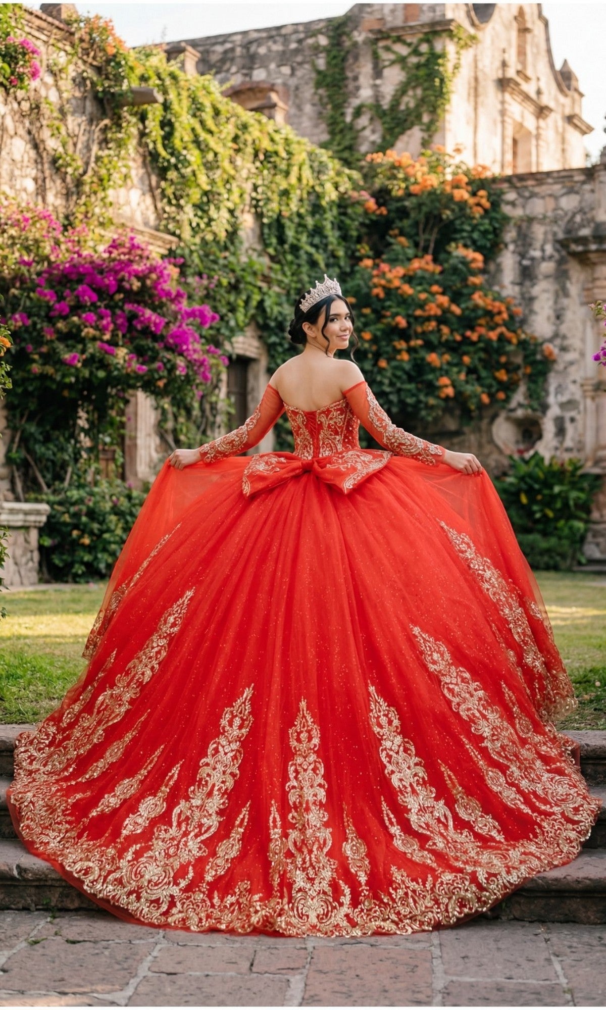 A woman in the Quinceanera Dress 8545JQ By Cinderella Couture, featuring red and gold hues with beaded sequin embellishments and an off-the-shoulder sweetheart neckline, stands on stone steps surrounded by flowers and garden walls.